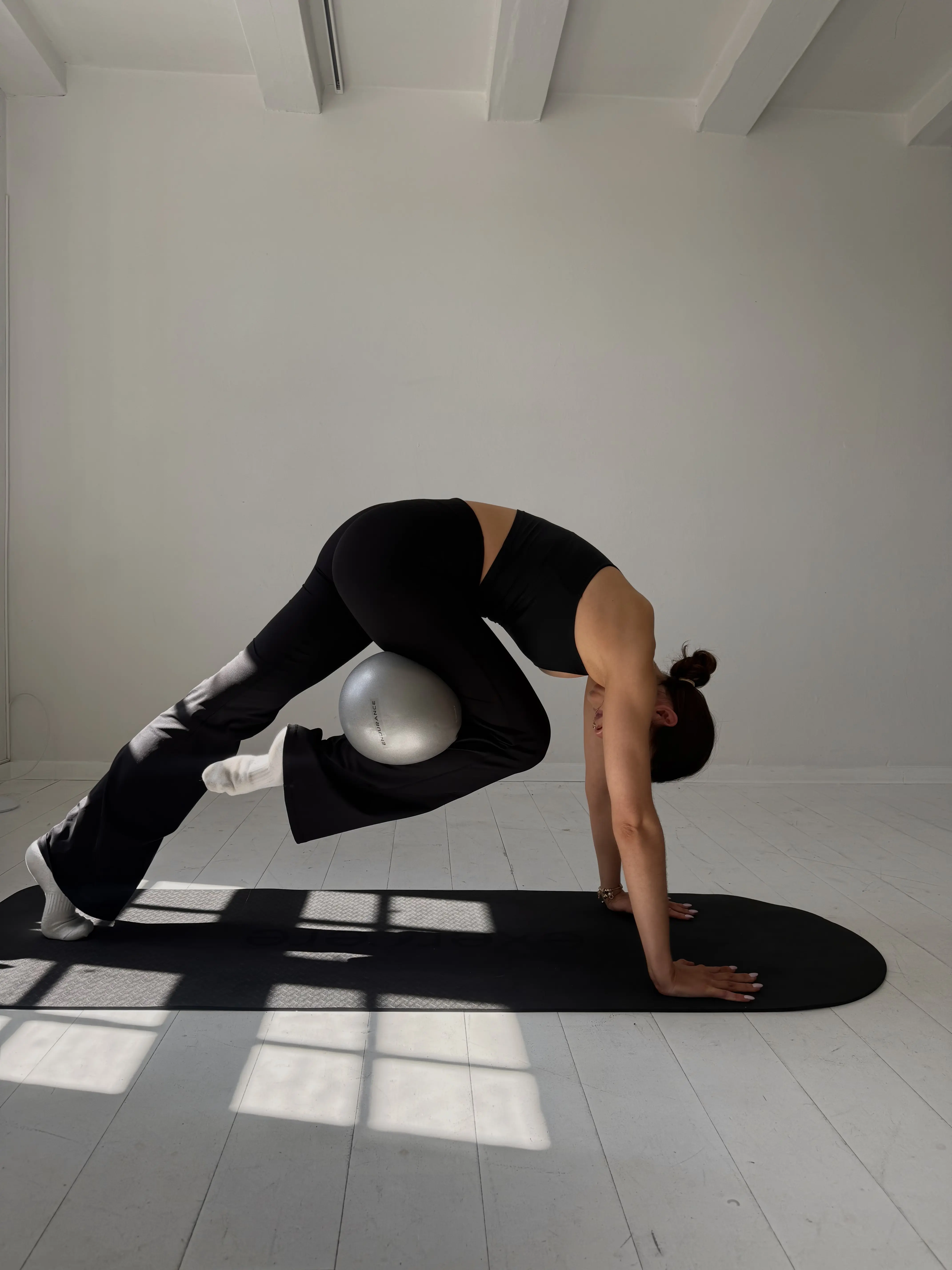 A woman practising Pilates with a small exercise ball on a mat in a sunlit studio.